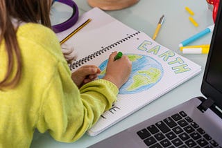 A child wearing a yellow sweater is drawing the planet Earth on a notebook. The word “Earth” is colored in green.
