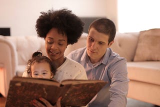 Interracial heterosexual couple of white male and brown-skinned female with short curly hair. The mom is holding a 2 year old on her lap and they’re reading her a story from a big, brown, old-looking book. They are sitting on a living room floor next to the couch.