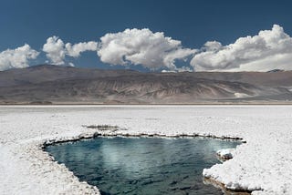 a spring of water in a desert, possibly salt, blue sky and white clouds