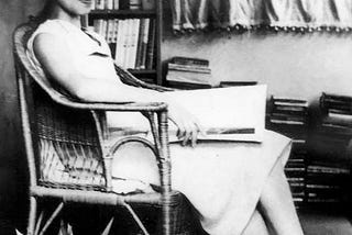 Sagawa sitting in a chair in a white dress with a book on her lap, her hair in a bun, wearing glasses.