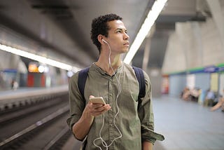 A young man walks through a transit station while listening to music through wired headphones.