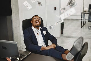 Man in a suit sitting at a desk with his feet up, smiling joyfully as hundred-dollar bills fall around him in an office setting.