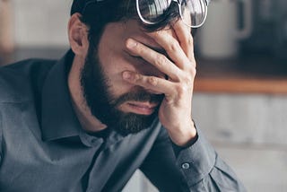 A bearded man with eyes shut holding his face in his hands.