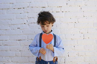 small boy holding a red, paper heart, standing in front of a white brick wall