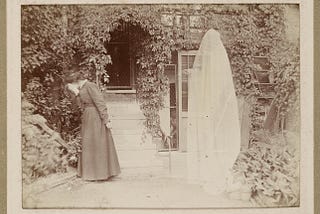 In a lush courtyard, close to a glazed and overgrown building entrance, a woman covers her face and turns away from a white-translucent sheet-covered ghost pointing at her with its singularly visible hand.