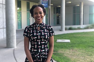 Brittnay Proctor stands outside of Humanities Gateway smiling and holding her hands in front of her.