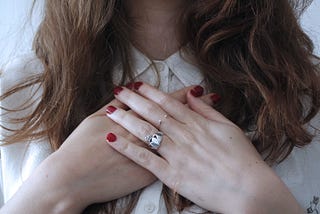 A brunette, white woman with her hands with red painted nails and a rings on her heart space