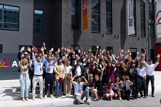 Group photo of approximately 50 adults of different genders and nationalities posing outside of a university building with their hands in the air and cheering.