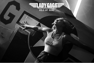 Lady Gaga Hold My Hand cover image: Lady Gaga in a white tank top and dog tags spreading her arms against the wing of a fighter plane