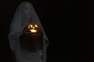 a spooky dark picture of a young girl in a ghost costume holding a lit jack-o-lantern