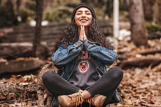 Smiling woman sitting in forest in jean jacket and hiking boots, hands in prayer posture under chin.