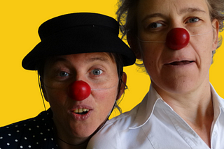 A bright yellow background, and the two sacred clowns, Katrijn and Ida, with red noses and expressive faces looking towards the viewer.