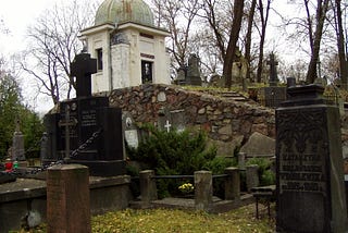 Foreground is individual graves with headstones. In background is a knoll with a small mausoleum with more individual headstones beyond it.