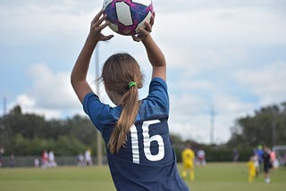 Photo of a girl throwing a soccer ball. It is shot from the back and other players are out of focus.
