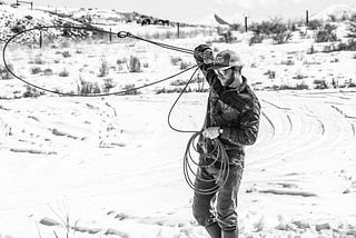 Black and white photo of man twirling a lasso in snow-covered Wyoming.