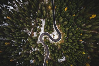 A helicopter view of a curved mountain road surrounded by green pine trees and a slight bit of snow.