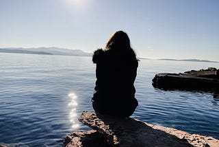 A woman sits in fetal position on a rock ledge beside a lake.