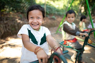 A young Thai girl is wearing a green and white shirt and playing on a playground with several other children.
