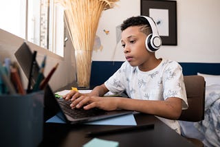 A young Black boy works on a laptop while wearing headphones. Photo by Vergani_Fotografia/Getty Images