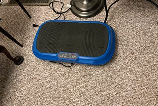 Blue LifePro vibration plate exercise machine with a black textured surface and control panel, placed on beige carpet near a lamp and ventilation grate.