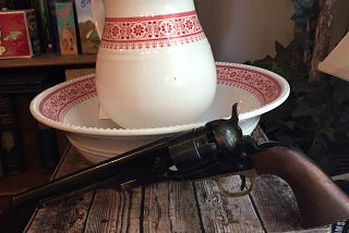 An old-fashioned ceramic pitcher and matching wash basin with matching red floral patterns, and an old cap and ball Colt revolver, displayed on a weathered wood crate.