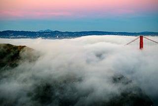 Foreground of fog in turmoil with tip of San Francisco Bridge showing above it. Background of rosy morning light on horizon.