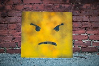 A canvas with a yellow angry face painted on it rests against a brick wall.