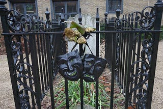 The grave of Joseph Grimaldi. A weathered headstone and grave is surrounded by black, ornate, cast-iron railings. The twin masks of tragedy and comedy — also in black iron — hang on the railing.