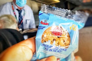 A photo from the first class seat of an airplane, author holding up a cookie in a package near with the Steward in the background. There’s an image of a little man on the cookie wrapper that looks uncannily like the Steward