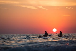 Two people sit on paddle boards in the ocean as the sun sets behind them