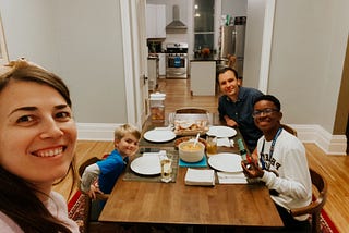 World Learning’s Youth Ambassador Brayan Weelkly Prendigan is sitting with his host family at a square dining room table. He is next to the father, a young boy is across from him, and a mother is taking the selfie shot.