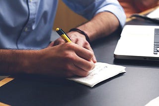 A person writing on paper next to a laptop