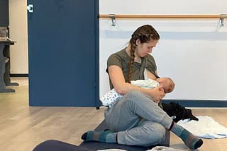 A woman on a yoga mat sits in cow face pose while nursing a baby