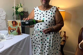 CaspHer’s mother in a pretty white dotted dress. She is holding a champaign bottle and glass. Behind her is a photo of herself years ago doing the same pose. Mum wears red lipstick and is smiling peacefully in your direction.