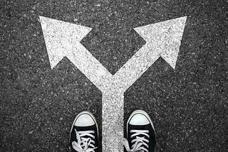 Photo of a pair of black and white sneakers on a pavement. Ahead, 2 white painted arrows point towards the top left and top right.