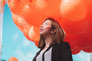 A woman in a business suit looks up at a net full of balloons above her. An excited/optimistic smile is on her face.