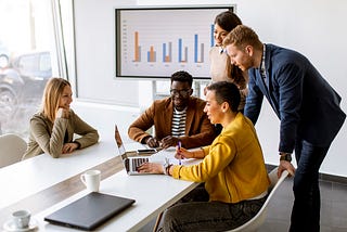 Five people sitting and standing around a conference table looking at a laptop.