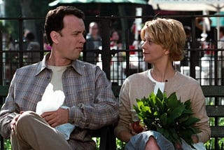 Tom Hanks and Meg Ryan as Joe and Kathleen in You’ve Got Mail sit looking at each other on a bench in New York.