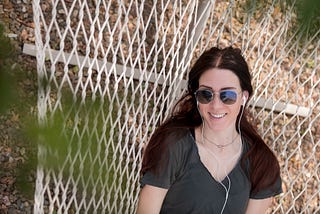 A twenty-something’ woman, with earbuds, smiles while lying in a hammock and looking up through tree branches.