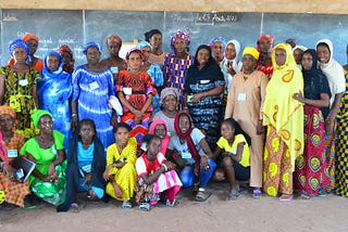 Three generations of women, wearing brightly colored outfits and standing in front of a chalkboard, who participated in an All Women’s Forum, Saré Konco village.