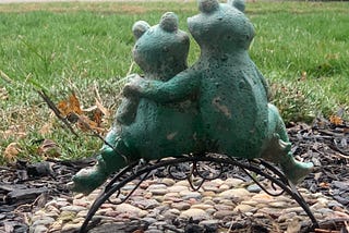 A statue of two frogs sitting beside each other, one with their hand around the other’s shoulder