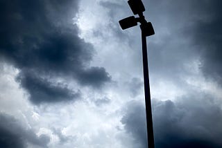 A silhouette of a parking lot streetlight with three bulbs, with a background of dark ominous clouds