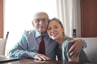 Sitting and smiling elderly man with left arm around seated and smiling younger woman who is a family member
