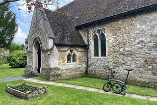 A Brompton bicycle sitting on a path outside an old stone church. The sky is blue. The grass is green. There is a little raised flower bed in the foreground. The flowers have yet to bloom to their full potential.