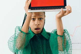 A professional Black Woman is holding an open laptop over her head. In teh computer screen, we can read the words “SCAM”