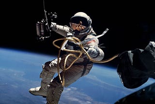 An American astronaut outside of his spacecraft, with a tether attaching his suit to the capsule, and the horizon of Earth and outer space in the background.