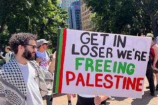 A Palestinian protestor holding a sign stating, “Get In Loser We’re Freeing Palestine”
