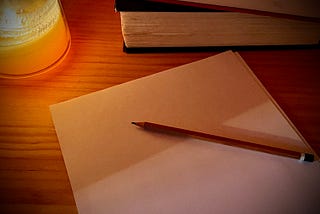 Some sheets of blank paper on a wooden surface. There is a wooden pencil on the papers, a stack of books in the background, and there is a lit candle providing a soft glow in the corner.