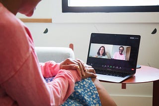 A woman conducts a teletherapy session on a laptop. The screen of her computer shows two people in a Zoom call.