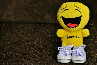 A bright yellow plush toy with a large, joyful laughing face stands on a dark textured surface. The toy has “Happy…” embroidered on its body and is wearing small white lace-up sneakers.